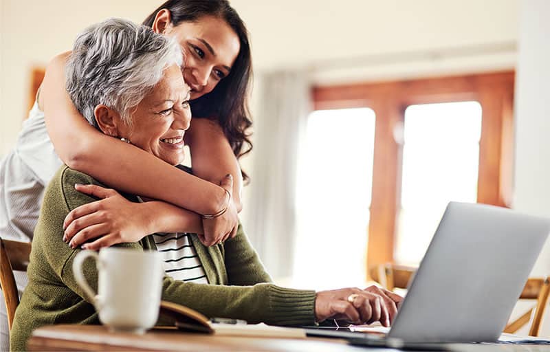 A woman hugging her mom while she types on a laptop